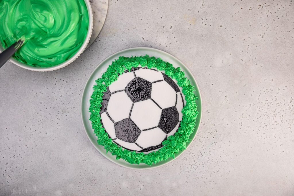 The completed soccer ball ice cream cake rests in the center of the image, while the large bowl of green icing and spoon remain in the top left corner.
