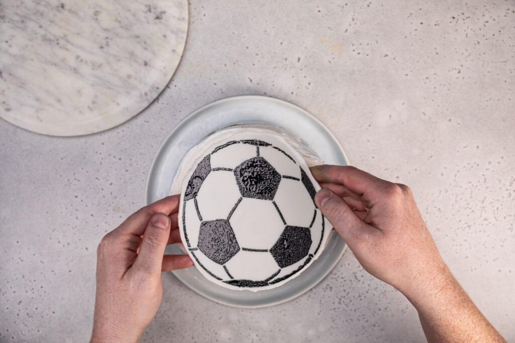 Dos manos colocan el fondant con el patrón de balón de fútbol en la parte superior del pastel de helado.