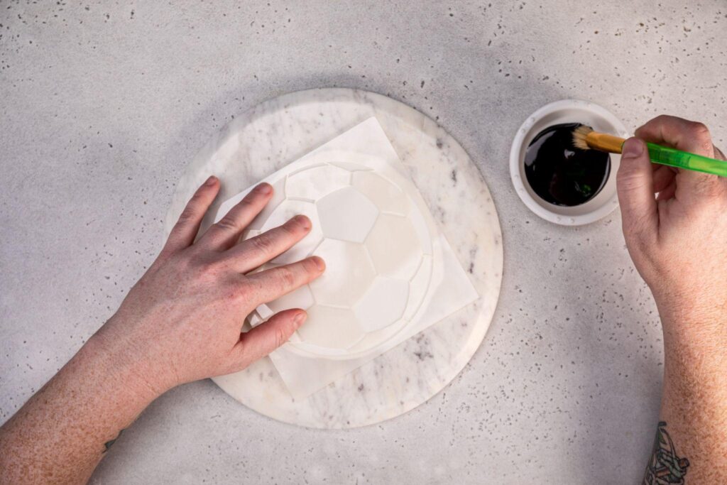 The soccer ball stencil rests on top of the fondant, which sits on a circular stone plate that looks like marble. A hand on the left holds the stencil, while the right hand dabs the paintbrush into the food coloring.