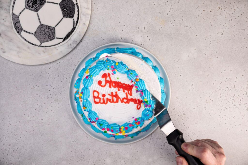 El fondant con el patrón de balón de fútbol se ha movido a la esquina superior izquierda de la imagen. En el centro, la mano de una persona utiliza un cuchillo para glaseado para retirar la glaseado congelado de la parte superior de un pastel de helado Carvel Happy Birthday.