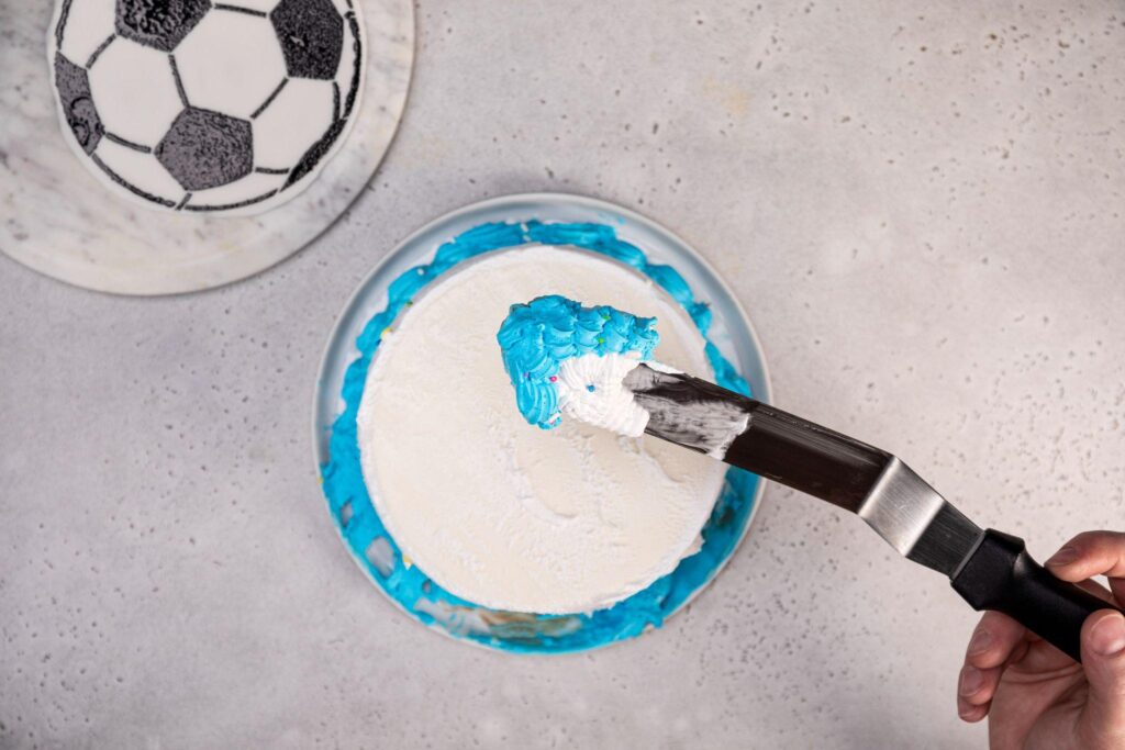 El fondant con el patrón de balón de fútbol permanece en la esquina superior izquierda de la imagen. Un pastel de helado Carvel Happy Birthday sin glaseado en la parte superior se encuentra en el centro de la imagen.