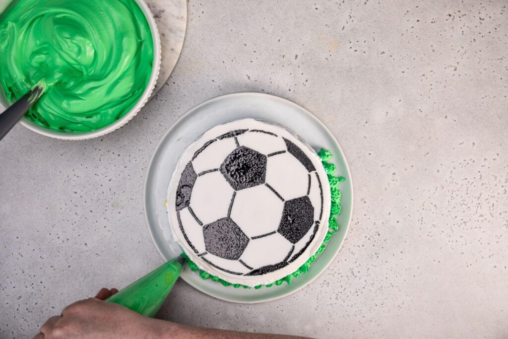 A large bowl of green icing with a spoon in it rests on the stone plate in the top left corner. The soccer ball cake is in the middle, and the person’s hands add icing to the bottom of the ice cream cake.